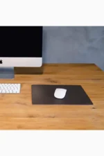 Dark brown leather mouse pad placed on a natural wood desk, with a white wireless mouse in the center, next to a desktop monitor and a white wireless keyboard, against a grey concrete wall background.