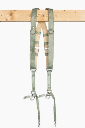 Full-length front view of the Green Marble leather dual camera harness hanging from the "Genuine Strap" branded wooden beam, showing the complete harness with cream leather crossback, marbled sage-green straps, silver buckles, D-rings, triangular connectors, and snap-hook camera attachments with green leather safety tabs at the bottom.
