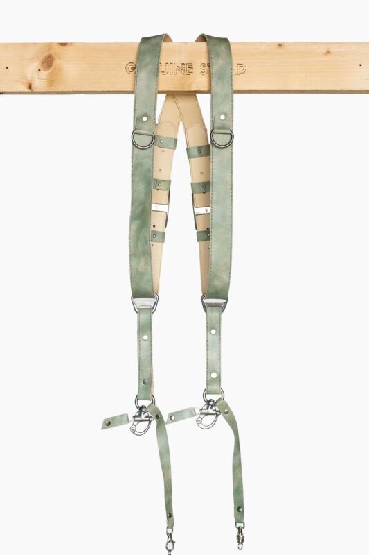 Full-length front view of the Green Marble leather dual camera harness hanging from the "Genuine Strap" branded wooden beam, showing the complete harness with cream leather crossback, marbled sage-green straps, silver buckles, D-rings, triangular connectors, and snap-hook camera attachments with green leather safety tabs at the bottom.