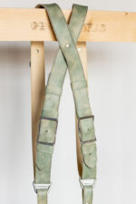 Close-up of the X-shaped crossback junction of the Green Marble harness hanging from a "Genuine Strap" branded wooden beam, showing the marbled sage-green leather crossing with a center rivet, dual silver square buckles, and matching green leather keeper loops.