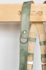 Detail of a single shoulder strap draped over the "Genuine Strap" branded wooden beam, highlighting the marbled sage-green leather texture, a riveted D-ring attachment loop, and the cream leather backing with green keeper loops visible in the background.