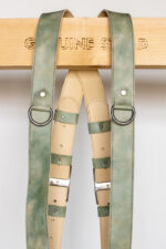 Front-facing view of the upper portion of both straps hanging from the "Genuine Strap" branded wooden beam, showing the marbled sage-green leather shoulder straps with riveted D-rings, cream leather padding with stitched rounded tabs, green keeper loops, and eagle-embossed silver slide buckles.