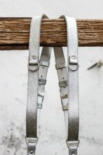 Grey Flower Embossed leather camera harness displayed on weathered wood beam, showing full X-harness design with silver-grey shoulder straps featuring embossed floral pattern, light grey center strap with embossed flower design and darker grey accent bands, metal D-ring connectors with ombre color gradient