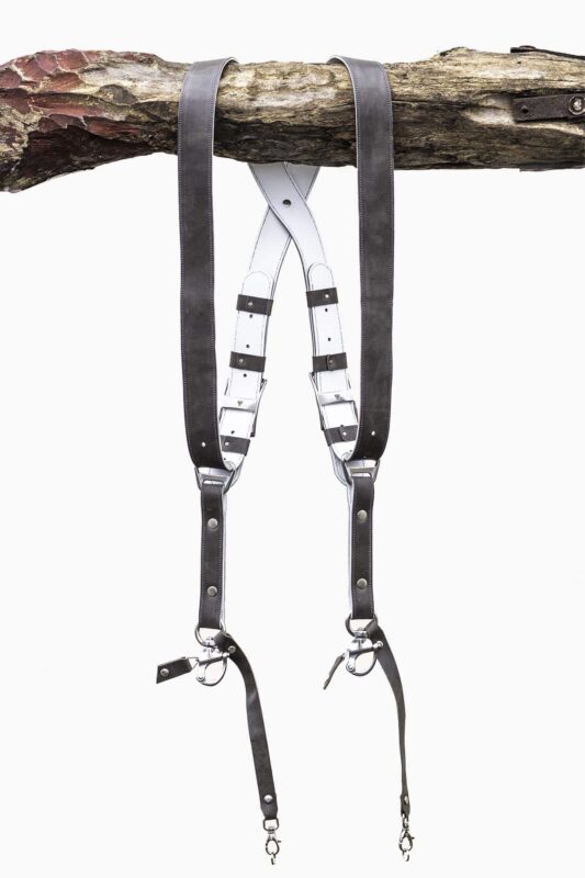 Grey goose vintage leather dual camera harness hanging from a rustic wooden branch against a white background, showing the full front view with two shoulder straps, a white leather back panel with leather keepers, adjustable silver buckles, riveted construction, and snap shackle clips with lobster claw clasps at each end.