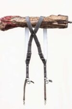Full view of the grey goose vintage leather dual camera harness hanging from a rustic wooden branch, displayed from the back showing the X-cross strap configuration, white leather backing, silver adjustable buckles, and dual snap shackle attachment points with leather safety tethers at the bottom.