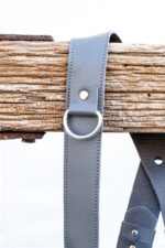 Close-up of Grey Sandstone harness shoulder strap showing stacked leather segments, metal ring connector, and leather attachment point on weathered wood