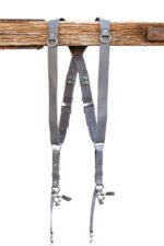 Grey Sandstone leather camera harness displayed on wooden beam showing full X-harness design with dual shoulder straps, adjustable leather segments, and metal D-rings