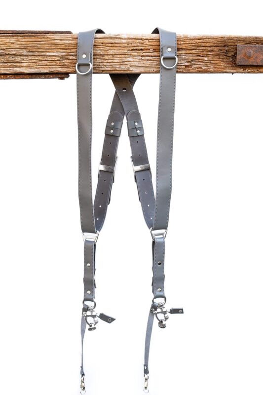 Grey Sandstone leather camera harness displayed on wooden beam showing full X-harness design with dual shoulder straps, adjustable leather segments, and metal D-rings