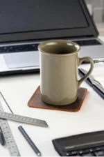 A dark brown leather coaster with contrast stitching on a white desk, with an olive-green ceramic mug placed on top of it, surrounded by office supplies including a ruler, pens, and a laptop in the background.