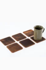 Six square brown leather coasters with contrast stitching laid out in a 2×3 grid on a white surface, with an olive-green ceramic mug sitting on the top-right coaster.