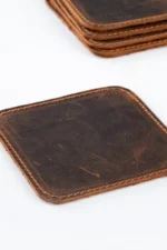 Close-up of a single square dark brown distressed leather coaster with visible natural grain, scratches, and tan saddle stitching along the edges, with additional stacked coasters partially visible in the upper corner.