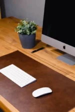 Top-down close-up of two leather desk mats side by side — one glossy medium brown and one matte dark brown — with a white wireless mouse resting on the darker mat, on a light stone surface.