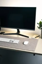 A light gray leather desk mat on a white desk with a white wireless keyboard and mouse on top, a widescreen monitor behind, a small yellow notepad, a pen, and a potted plant to the right.