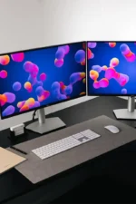 A dark gray leather desk mat on a black desk beneath a dual-monitor setup displaying colorful abstract wallpaper, with a white wireless keyboard and mouse, a pen, and a notebook on the side.