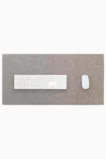Top-down view of a light gray leather desk mat with stitched edges on a white background, with a white full-size wireless keyboard centered on the left and a white wireless mouse on the right.