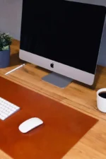 A tan cognac leather desk mat on a natural oak desk with a white wireless keyboard, a white wireless mouse, a white pen, and a coffee cup in a ribbed white mug, with an iMac monitor in the background and a small potted plant.