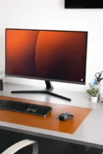 A tan cognac leather desk mat on a light gray desk with a black wireless keyboard and black mouse, under a widescreen monitor displaying a warm orange-brown gradient wallpaper, with desk accessories and a small plant to the right.