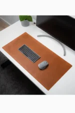 Overhead angled view of a tan cognac leather desk mat on a white desk, with a compact dark keyboard and a gray wireless mouse on top, a potted plant in the corner, and a curved monitor stand visible at the edge.