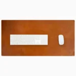 Top-down view of a rich cognac-tan leather desk mat with natural patina and subtle edge stitching, with a white full-size wireless keyboard and white wireless mouse placed on it, on a white background.