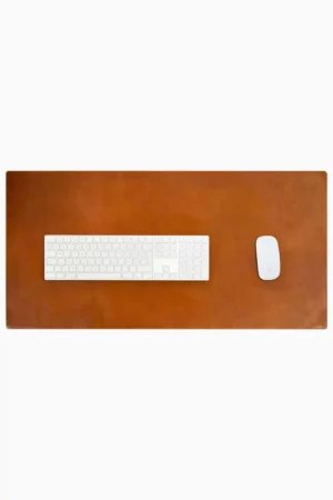 Top-down view of a rich cognac-tan leather desk mat with natural patina and subtle edge stitching, with a white full-size wireless keyboard and white wireless mouse placed on it, on a white background.