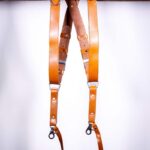Light Brown Garioch leather camera harness displayed flat on weathered wood showing full X-design with dual bright orange shoulder straps and brown center strap
