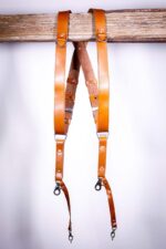 Light Brown Garioch leather camera harness displayed flat on weathered wood showing full X-design with dual bright orange shoulder straps and brown center strap