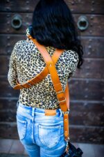 Close-up of woman wearing Light Brown Garioch harness in tan leather, holding professional DSLR camera, showing X-harness design and camera attachment