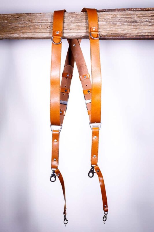 Light Brown Garioch leather camera harness displayed flat on weathered wood showing full X-design with dual bright orange shoulder straps and brown center strap