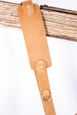 Light brown leather camera strap close-up with padded shoulder section, stitched edges, silver rivets, and metal ring detail hanging over a rustic wooden beam on a light gray background.