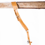 Full-length light brown leather camera strap with padded shoulder pad, adjustable strap holes, silver metal hardware, and clasp attachment hanging from a rustic wooden beam on a light gray background.
