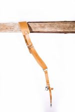 Full-length light brown leather camera strap with padded shoulder pad, adjustable strap holes, silver metal hardware, and clasp attachment hanging from a rustic wooden beam on a light gray background.