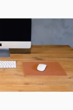 Light brown tan leather mouse pad placed on a natural wood desk, with a white wireless mouse in the center, next to a desktop monitor and a white wireless keyboard, against a grey concrete wall background.