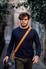 Man wearing the Garioch single camera strap in dark brown leather across the body, standing outdoors.