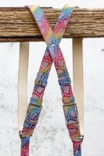 Both straps of the rainbow leather camera harness crossing in an X-shape below the rustic wooden beam, showing the natural leather padding behind, with silver square metal buckles at the bottom of each strap, against a grey textured background.