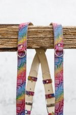 Upper section of the dual camera harness showing both shoulder straps hanging over a rustic wooden beam, with silver D-rings and rivets at the top, natural leather padding in the center, and adjustable buckle keepers with metal buckles below.