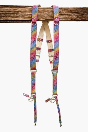 Rainbow multicolor leather dual camera harness with mosaic-textured snakeskin pattern, featuring natural vegetable-tanned leather padding, silver metal buckles, D-rings, and swivel snap hooks, hanging from a rustic wooden beam against a white background