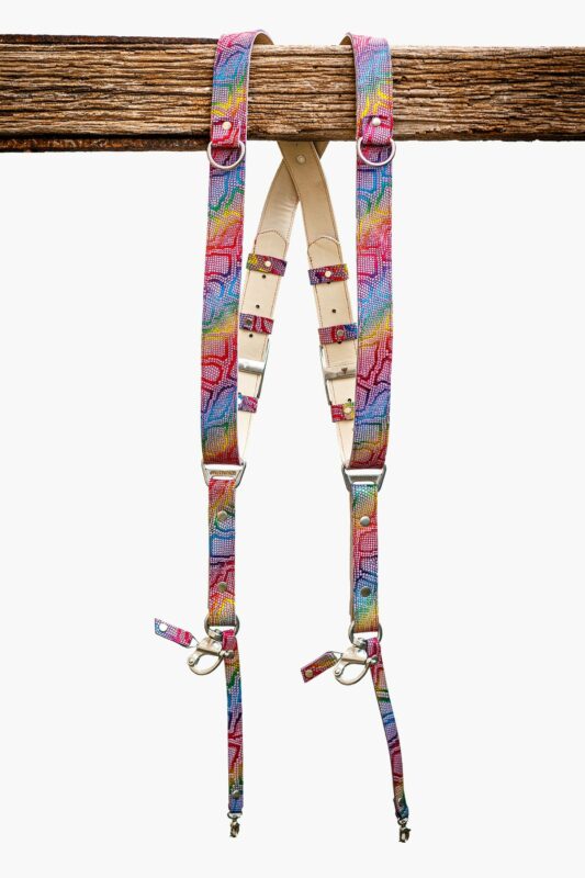 Rainbow multicolor leather dual camera harness with mosaic-textured snakeskin pattern, featuring natural vegetable-tanned leather padding, silver metal buckles, D-rings, and swivel snap hooks, hanging from a rustic wooden beam against a white background
