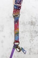 Close-up of the lower section of the rainbow leather camera strap, showing the silver metal triangular slider, rivets, swivel snap hook, and a small leather safety tab, against a grey textured background.