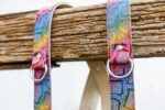 Close-up detail of the two shoulder straps draped over a rustic wooden beam, showing the rainbow mosaic leather texture with silver D-rings and rivets, with the natural leather lining partially visible underneath.