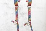 Detail of both lower camera attachment points of the rainbow leather harness, showing the branded metal sliders engraved with "GENUINESTRAP," silver rivets, O-rings, and swivel snap hooks, against a grey textured background.