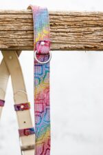 Side view of the right shoulder strap hanging over a rustic wooden beam, showing the full length of the rainbow mosaic leather with a silver D-ring, rivet, and the natural leather shoulder pad and buckle keepers visible in the background.
