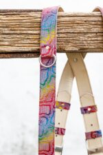 Side view of the left shoulder strap draped over a rustic wooden beam, displaying the full rainbow mosaic leather pattern with a silver D-ring and rivet, with the natural leather shoulder pad and buckle section visible behind it.
