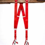 Red Campari leather camera harness front view showing shoulder straps draped over weathered wood bar, highlighting adjustable leather segments with metal connectors and center X-strap design