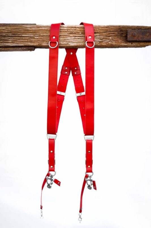 Red Campari leather camera harness front view showing shoulder straps draped over weathered wood bar, highlighting adjustable leather segments with metal connectors and center X-strap design