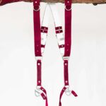 Red vintage leather dual camera harness with silver hardware, hanging from a rustic wooden branch against a white background, showing the full front view with two shoulder straps, a white leather back panel, adjustable buckles, and snap hook attachments at each end.