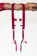 Red vintage leather dual camera harness with silver hardware, hanging from a rustic wooden branch against a white background, showing the full front view with two shoulder straps, a white leather back panel, adjustable buckles, and snap hook attachments at each end.