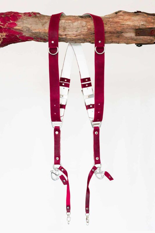 Red vintage leather dual camera harness with silver hardware, hanging from a rustic wooden branch against a white background, showing the full front view with two shoulder straps, a white leather back panel, adjustable buckles, and snap hook attachments at each end.