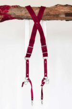 Full view of the red leather camera harness hanging from a rustic wooden branch, displayed from the back showing the X-cross strap configuration, white leather backing, silver buckles, and dual snap hook attachment points at the bottom.