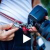 Close-up of hands attaching a DSLR camera to the red leather harness using a silver snap hook clip, showing the quick-release connection mechanism in action.