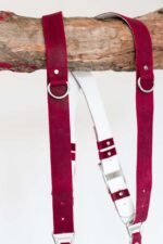 Close-up detail of the upper portion of the red leather camera harness hanging from a wooden branch, highlighting the silver D-rings, rivet construction, and the white leather backing with adjustable strap connections.